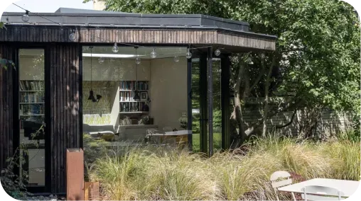Studio de jardin moderne en bois avec baies vitrées, intérieur bibliothèque visible, entouré de végétation