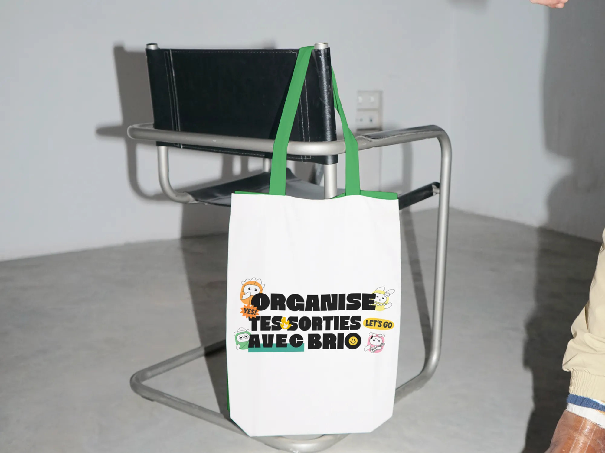 Tote bag blanc à anses vertes avec illustration "Organise tes sorties avec Brio", suspendu à une chaise design
