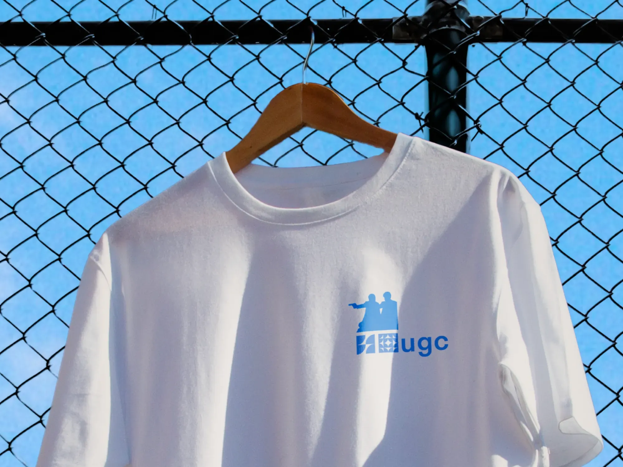 T-shirt blanc avec logo UGC bleu, suspendu sur un cintre en bois devant une clôture grillagée