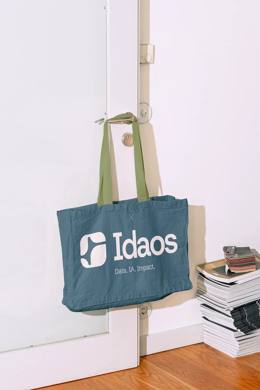 Tote bag bleu foncé avec logo Idaos "Data. IA. Impact." suspendu à une porte, pile de magazines en arrière-plan