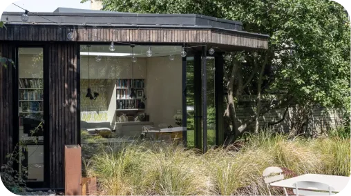 Studio de jardin moderne en bois avec baies vitrées, intérieur bibliothèque visible, entouré de végétation