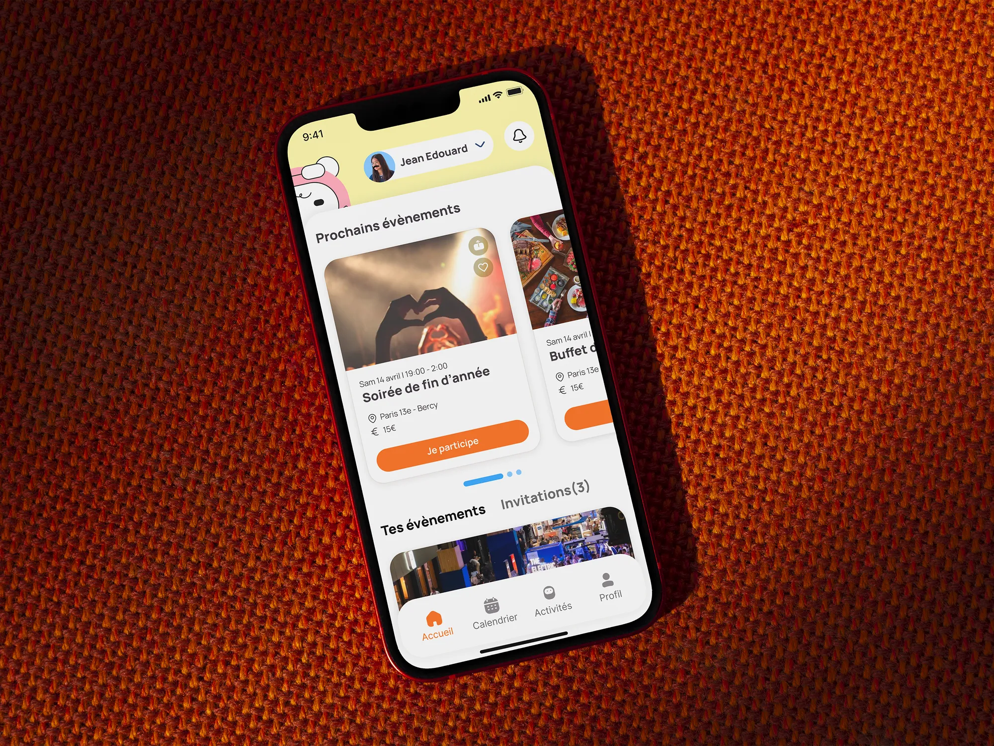 Smartphone posé sur textile orange affichant l'application Amigo, page d'accueil avec liste des prochains événements